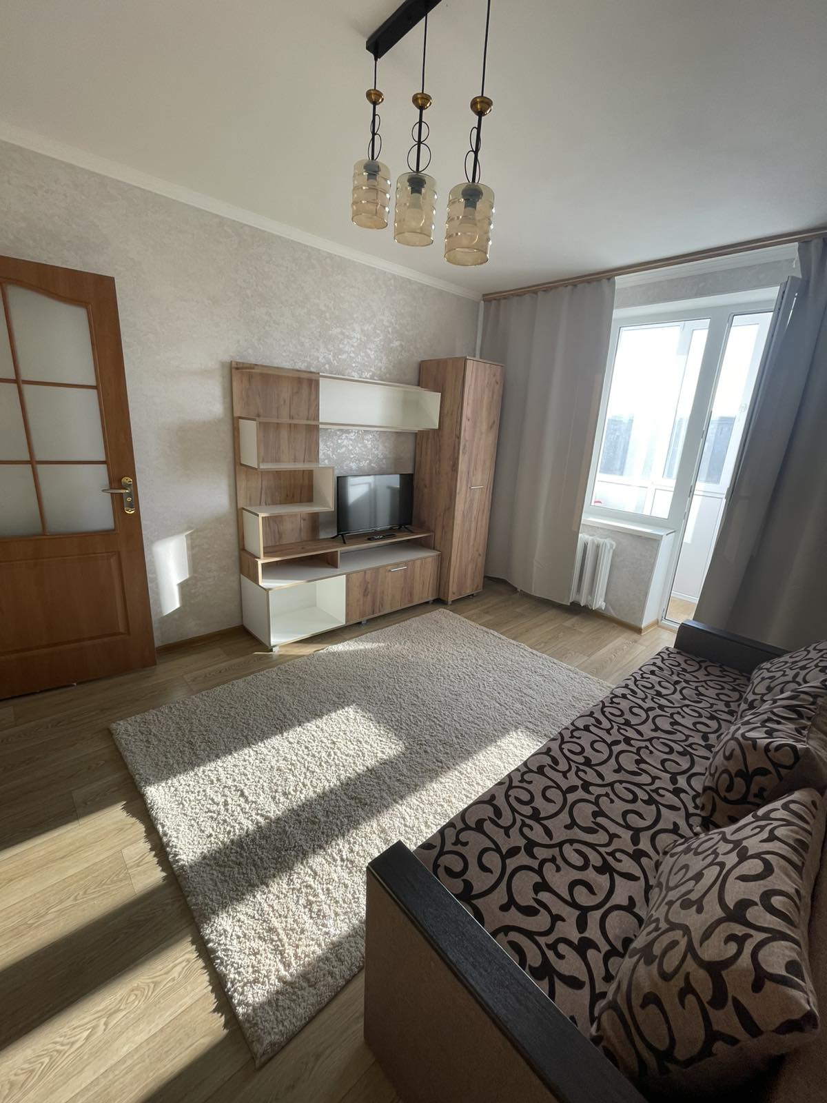 A light-filled living area features a patterned sofa with cushions, wooden shelving with a television, and a cozy area rug. Near the window, curtains gently filter sunlight, and a door leads to a separate room, creating an inviting and functional space.