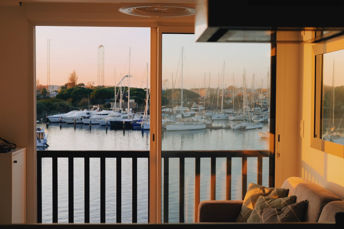 A serene view of a marina at sunset is captured through large glass doors. Boats are gently moored in the calm water, while warm light enhances the colors of the scene. A cozy seating area is visible in the foreground.