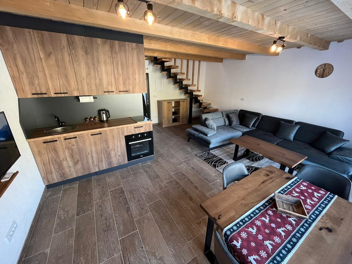 An open living space displays modern wood cabinetry and an equipped kitchen area. A spacious sectional sofa is located alongside a wooden dining table, which features a decorative table runner. A staircase to the upper level is visible in the background, complemented by wooden beams on the ceiling.