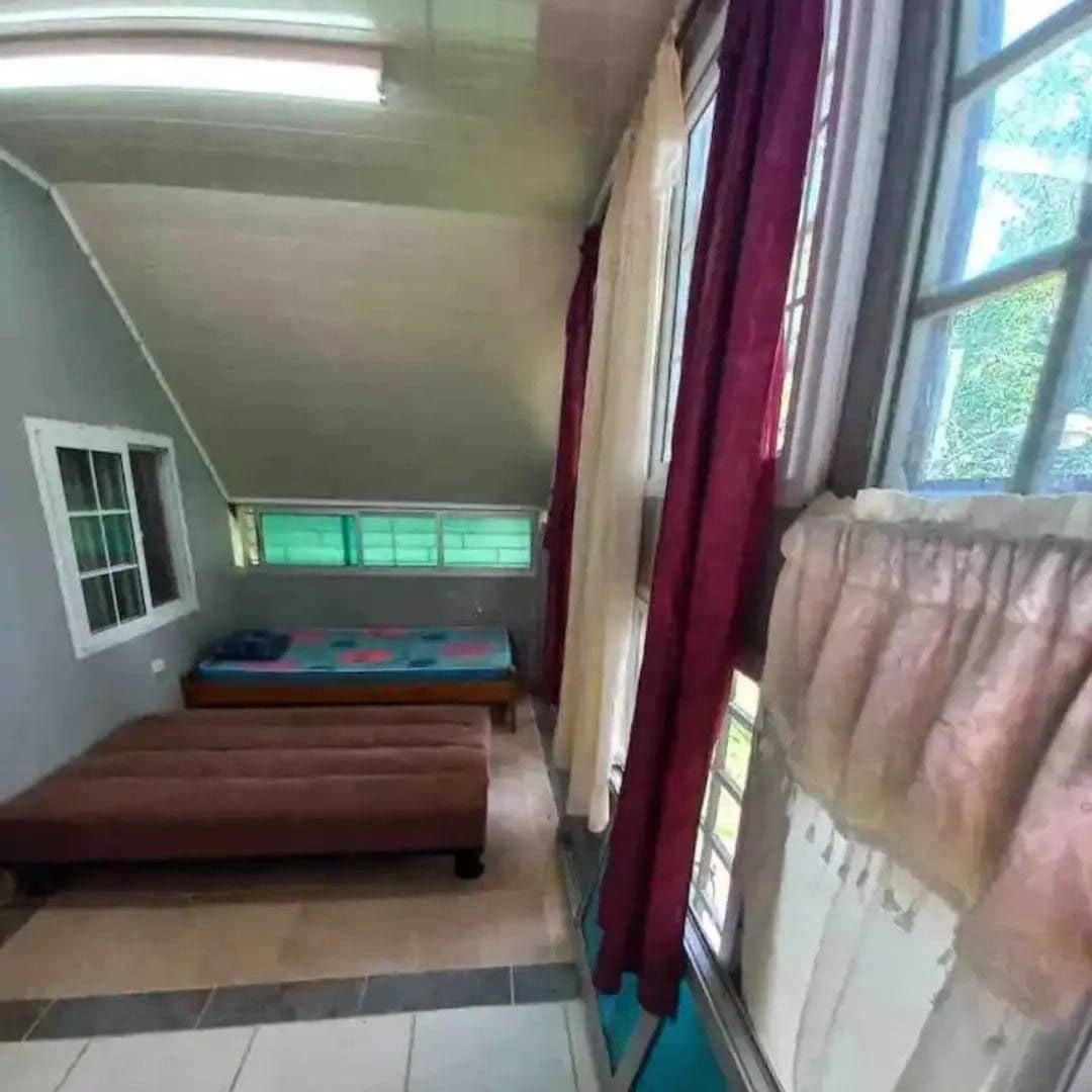 A spacious sleeping area features a neatly made bed and a cozy wooden bench. Large windows allow natural light to fill the room, while sheer curtains softly diffuse the sunlight, enhancing the tranquil atmosphere.