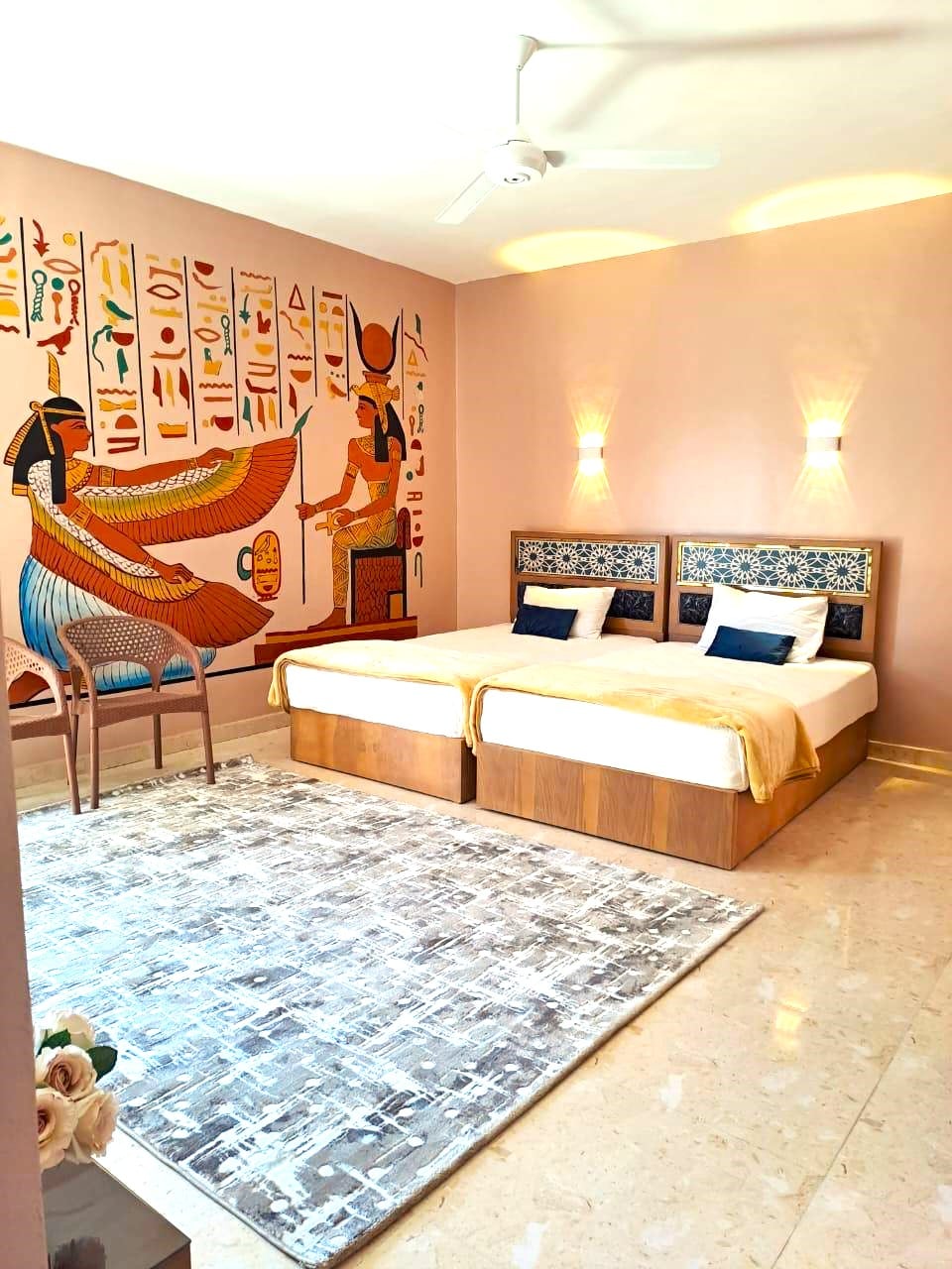 A bright sleeping area features two beds with decorative headboards. An artistic wall depicting ancient Egyptian motifs adds cultural charm. A chair is positioned beside the bed, and a plush rug provides comfort underfoot, enhancing the serene atmosphere of the room.