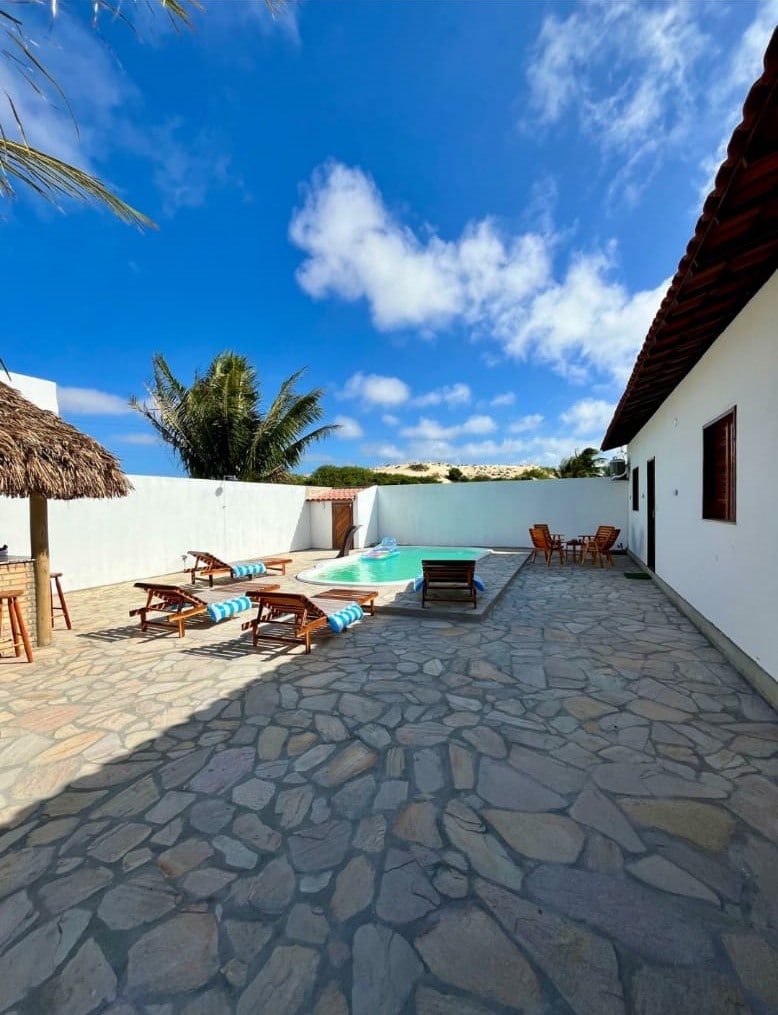 A serene outdoor area features a swimming pool surrounded by stone paving. Two lounge chairs are positioned near the pool, while a wooden table with chairs is visible on the patio. Lush palm trees enhance the inviting setting under a clear blue sky.