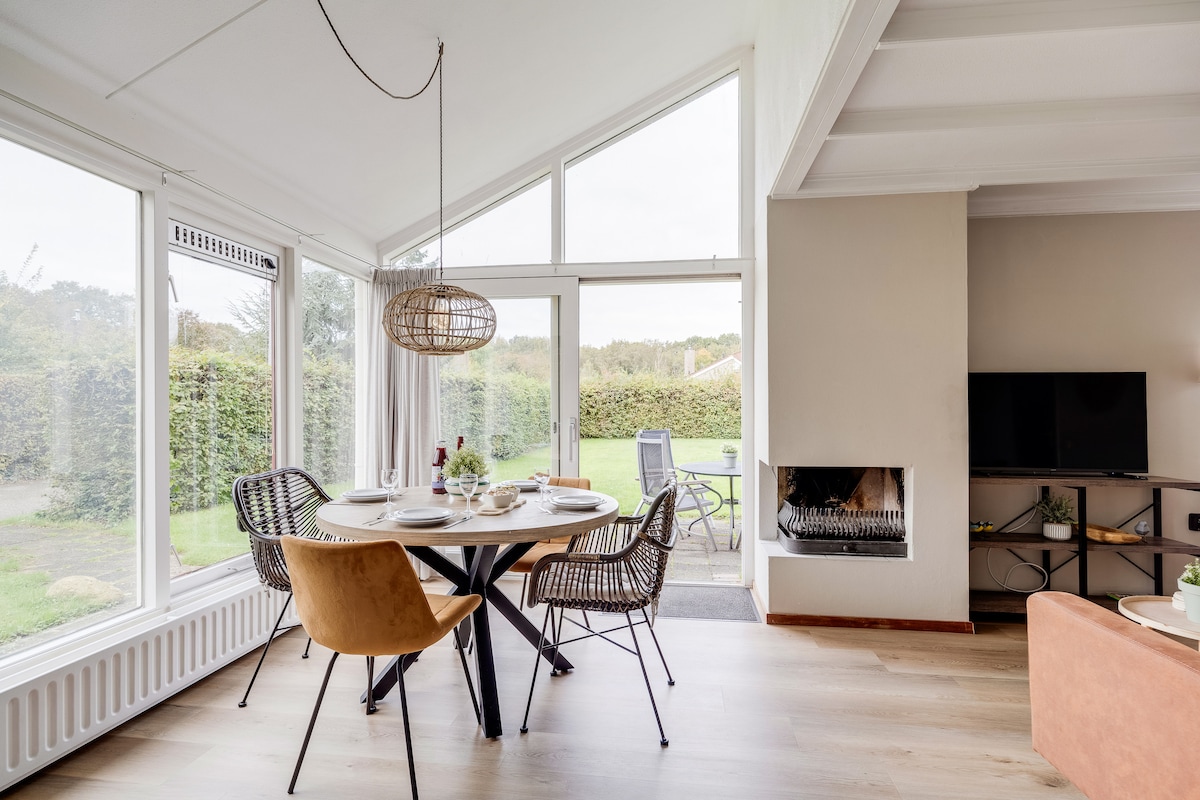 A living and dining area is illuminated by natural light streaming through large windows. A round table is set for dining, surrounded by modern chairs. A fireplace adds warmth to the space, while sliding doors provide access to the private garden outside.