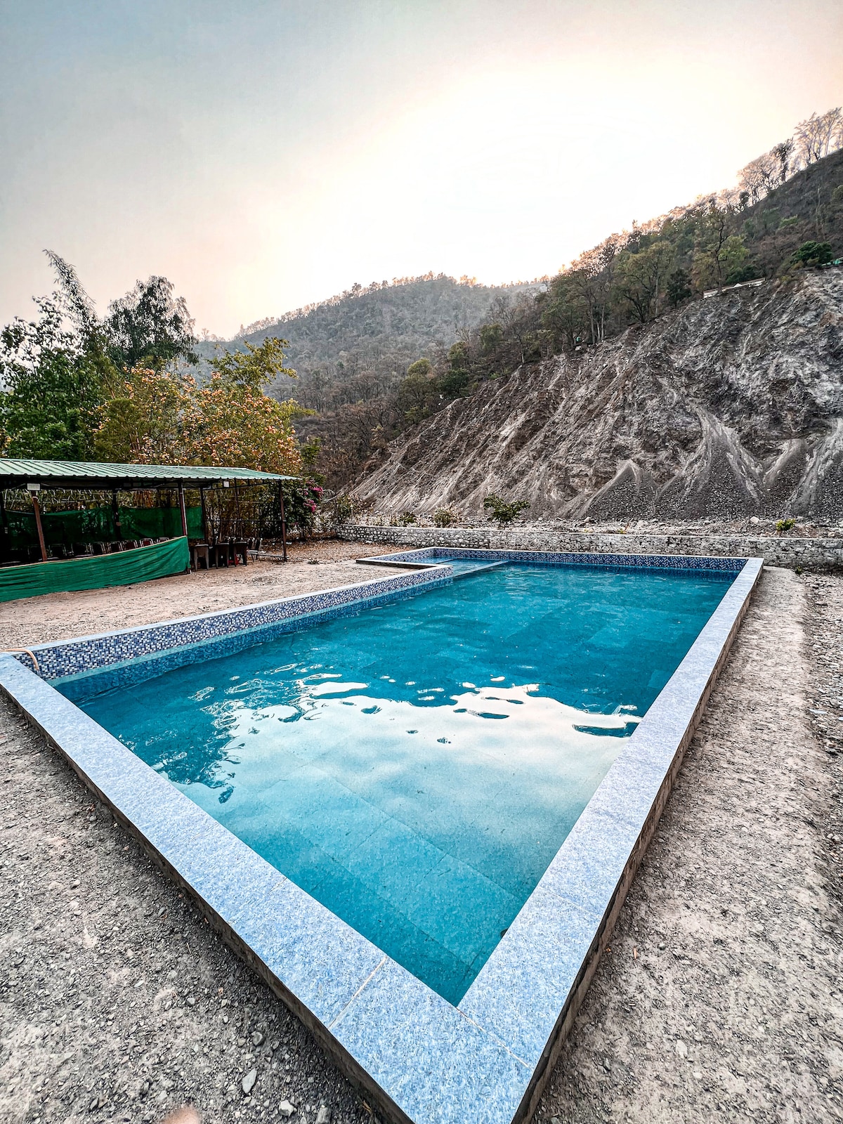 A rectangular swimming pool is situated amidst a natural landscape, surrounded by rocky hills and sparse vegetation. The clear water reflects the sky, while a shaded seating area is partially visible in the background, providing a place to relax by the pool.