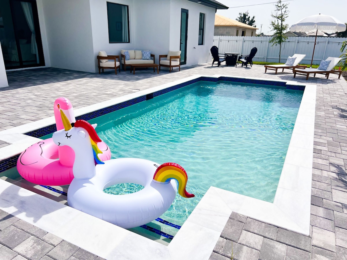 A private swimming pool is outlined with light-colored stone tiles, featuring colorful inflatable unicorns floating on the surface. Sun loungers and chairs are arranged around the pool, with a sun umbrella positioned nearby, providing shaded seating options.