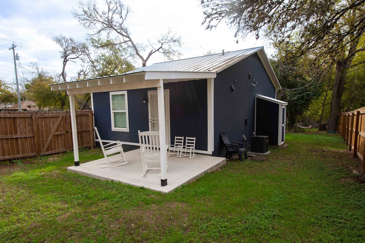 A covered patio area extends from the back of the house, featuring a small concrete slab with a couple of chairs white rocking chairs. The house has a dark blue exterior, surrounded by a green lawn and a wooden privacy fence.