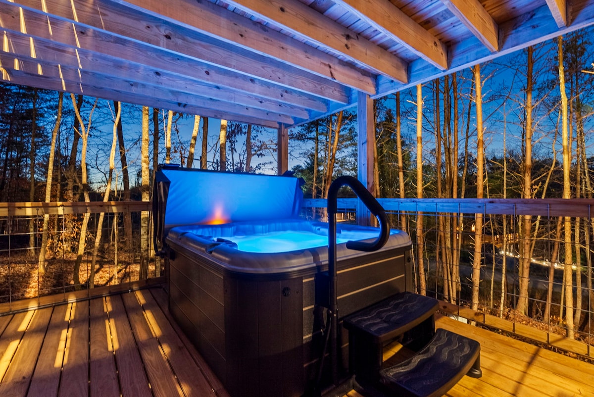 A hot tub is set on a wooden deck, surrounded by trees that create a natural canopy. Soft ambient lighting enhances the serene atmosphere, while the evening sky adds a calming backdrop. Steps lead to the tub, inviting relaxation and enjoyment.