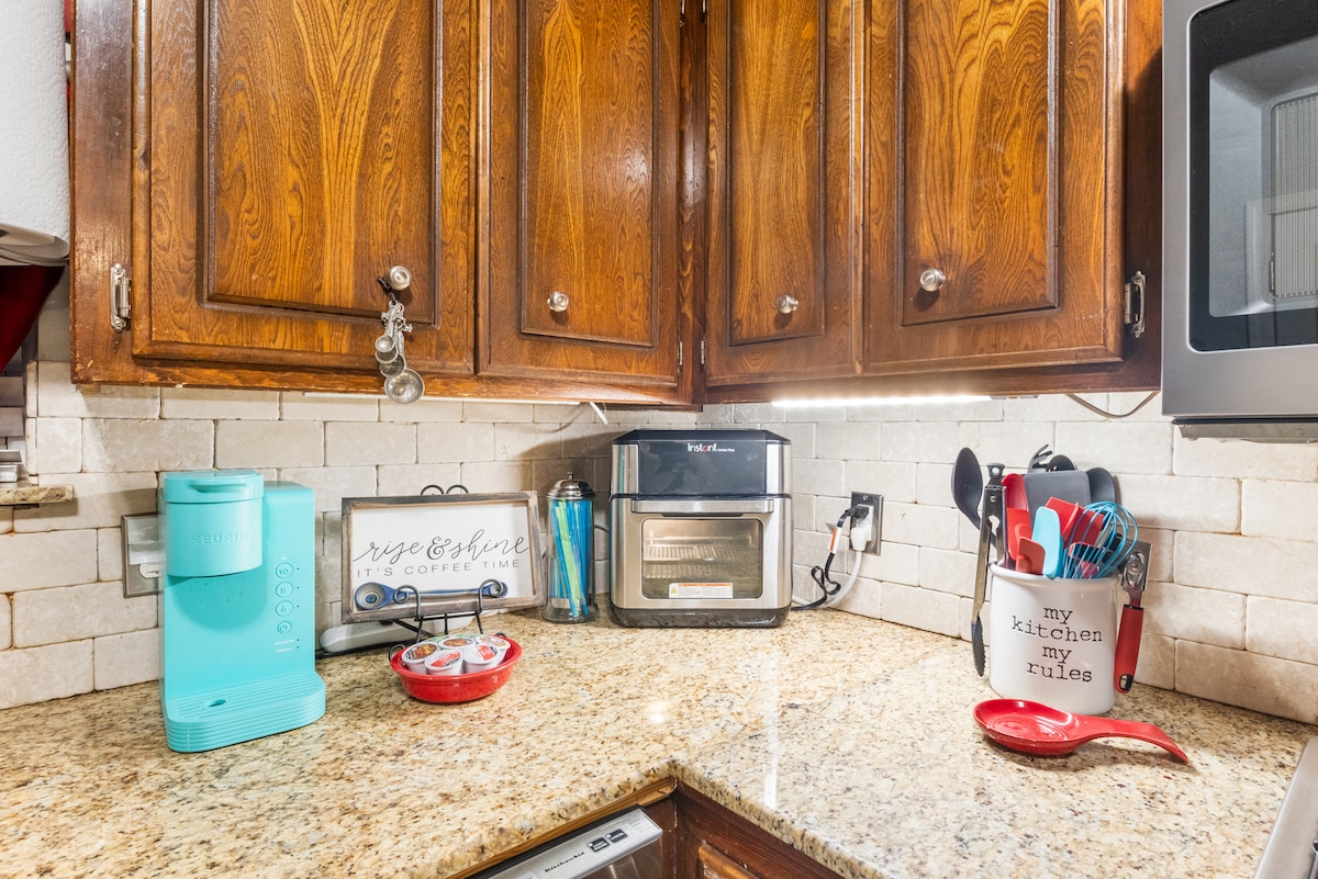 A well-equipped kitchen corner features dark wooden cabinets and a granite countertop. A turquoise coffee maker stands beside an air fryer, while cooking utensils are organized in a container. Bright lighting highlights the tidy arrangement, enhancing the kitchen's functionality.