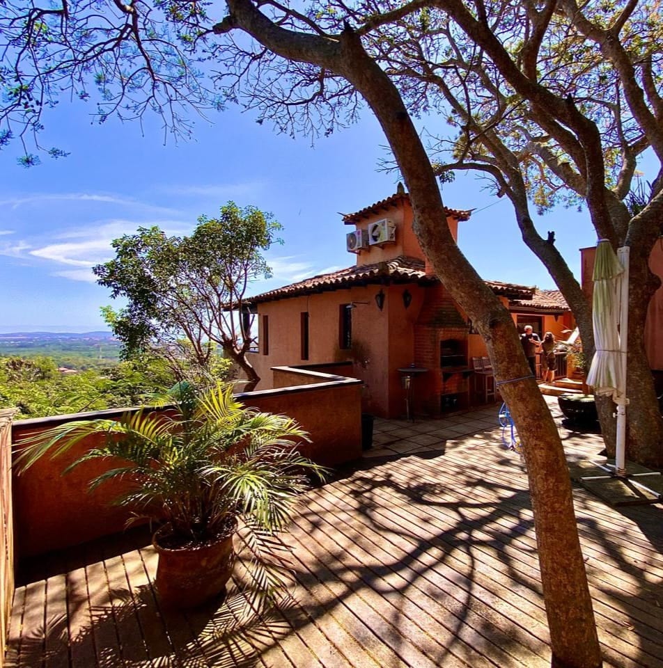 A spacious outdoor terrace is surrounded by trees, providing shade and a scenic view. A structure is visible in the background, featuring a tiled roof and an air conditioning unit. Potted plants add greenery to the setting, creating a relaxed outdoor environment.