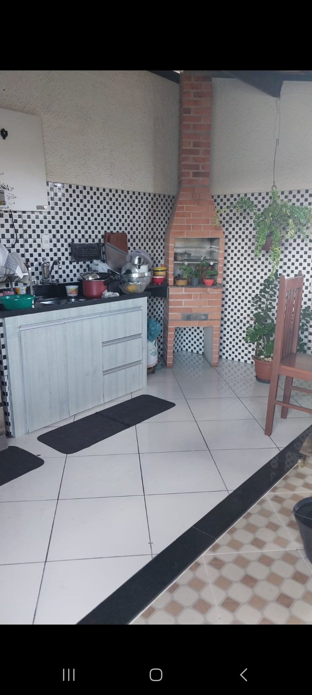 A kitchen area features a mix of black and white tiled walls, with a countertop holding various cooking utensils. A small brick barbecue stands against the wall, adorned with potted plants. Two chairs accompany a simple wooden table, adding functionality to the space.