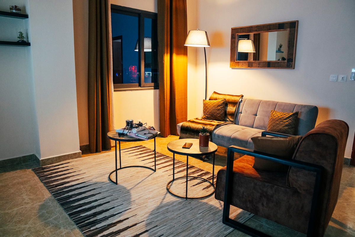 A modern living area is displayed with a plush sofa and accent chairs positioned around two round tables. A textured rug covers the floor, and warm ambient lighting is provided by a floor lamp. Large windows allow for evening views, framed by patterned curtains.