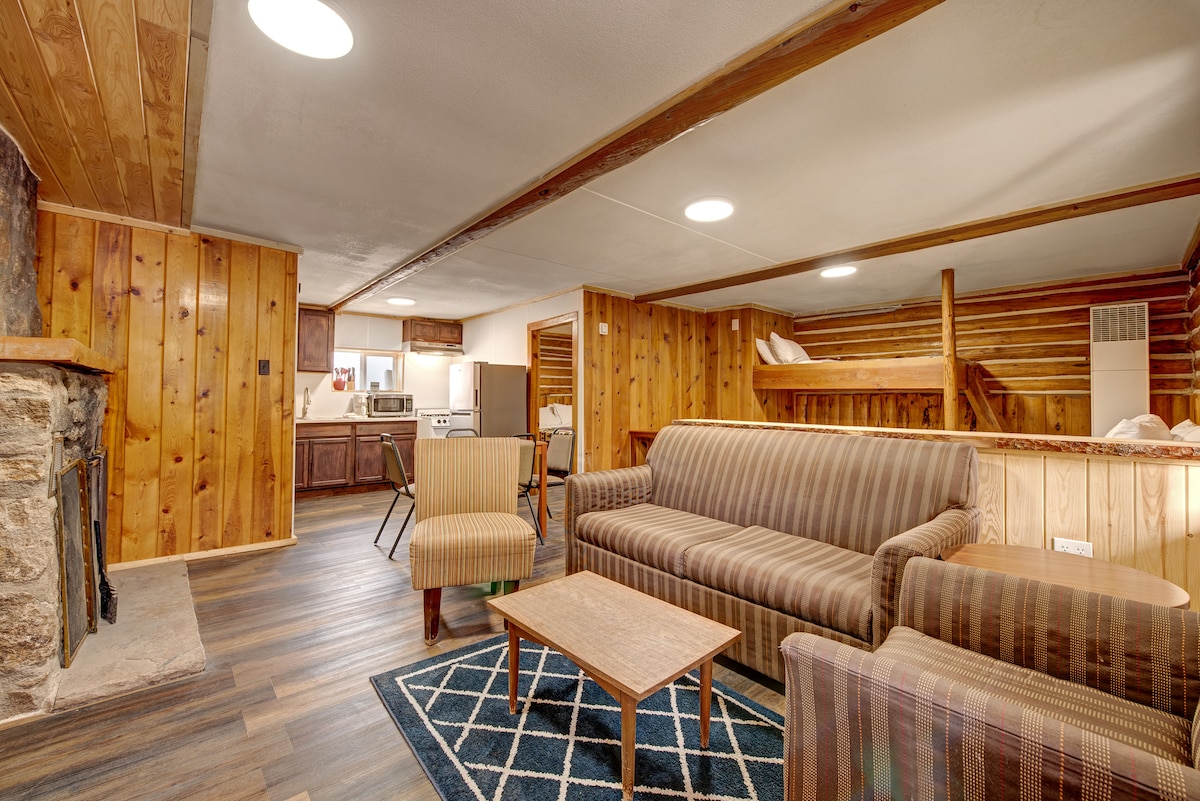 A welcoming cabin interior features wooden walls and a spacious layout. A striped sofa and an armchair surround a wooden coffee table on a patterned rug. A set of bunk beds is visible in the background, while a well-equipped kitchenette awaits in the corner.