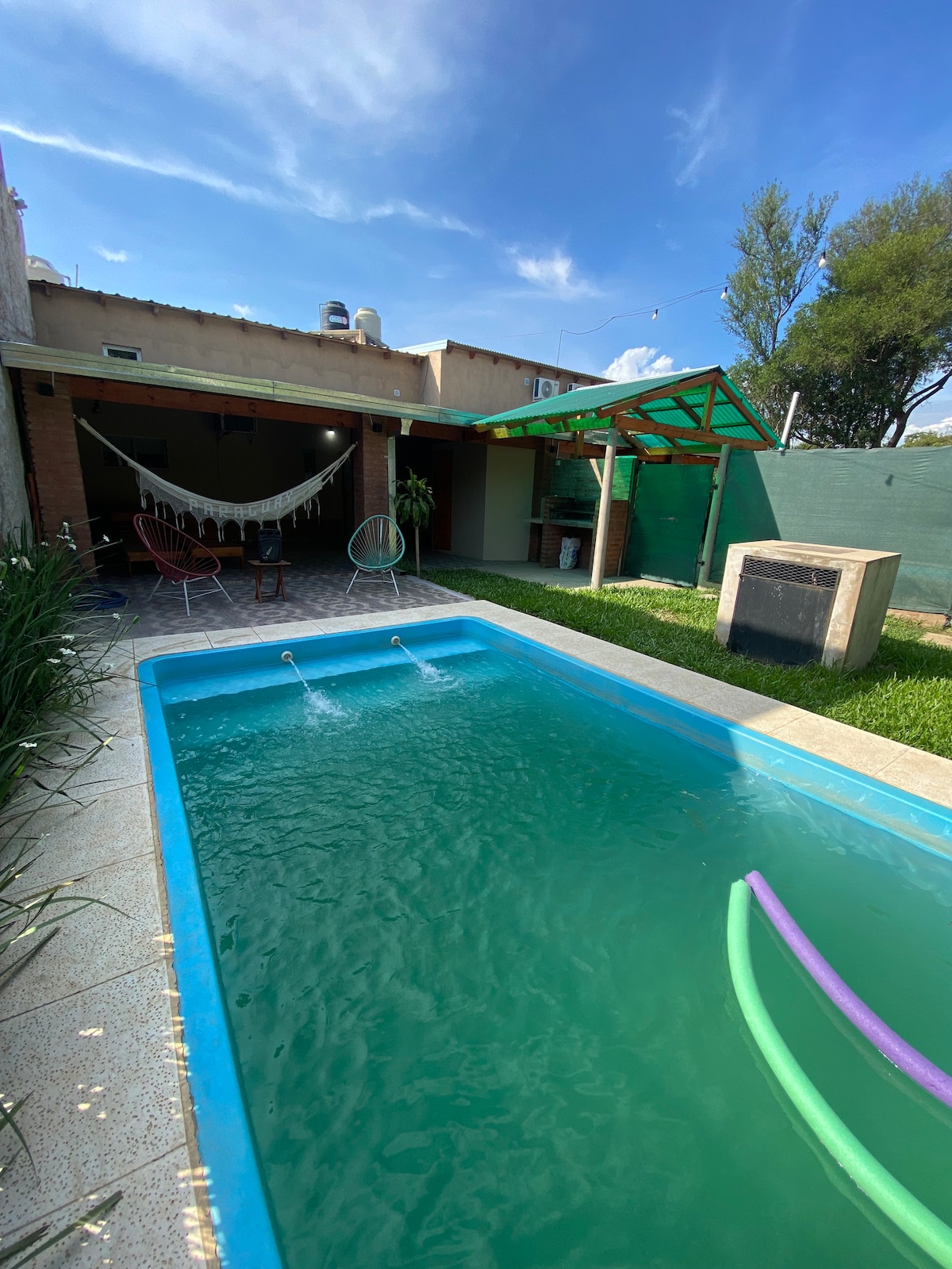 A refreshing pool with vibrant turquoise water occupies the center of the outdoor space, complemented by green lawn areas. A hammock is situated to one side, and colorful pool floats rest along the edge, enhancing the relaxing atmosphere of the backyard.