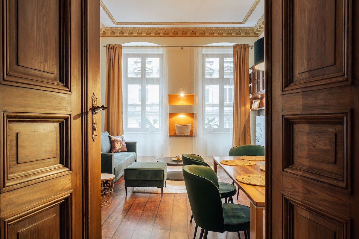 An inviting living area is visible through an open double door, featuring large windows with sheer curtains. A green velvet sofa, two patterned chairs, and a wooden dining table enhance the room's elegance. Decorative shelving is mounted on the wall, providing a modern touch.