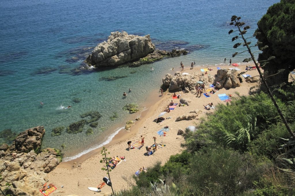A sandy beach is framed by rocky outcrops, with clear turquoise waters lapping at the shore. Sunbathers are seen on colorful towels, and small groups of people enjoy the water. The scenery includes lush greenery in the background, enhancing the vibrant coastal atmosphere.