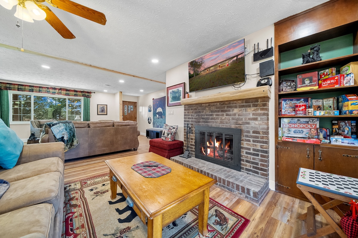 A welcoming living area is highlighted by a central fireplace surrounded by a brick mantle. Comfortable seating includes a large sectional sofa and a smaller chair. A wooden coffee table sits in front, while a bookshelf displays board games and decorative items. Natural light enters through the windows.