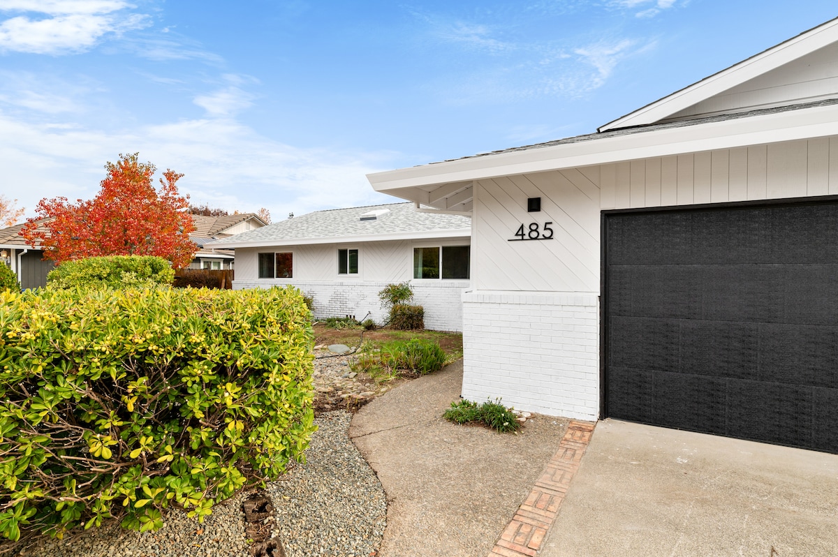 A modern home exterior is presented, featuring a clean white façade and a neatly manicured front yard. Lush green shrubbery frames the pathway leading to the entrance, and a spacious driveway leads to a dark garage door. Bright autumn foliage adds a touch of color.