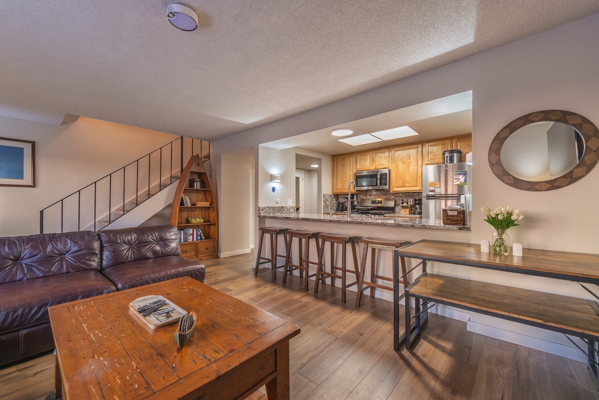The open living area features a large sectional sofa accompanied by a wooden coffee table. The adjacent kitchen, highlighted by wooden cabinetry, showcases modern appliances and stone countertops. A dining table with tall stools invites gathering, while natural light filters through the overhead skylight.