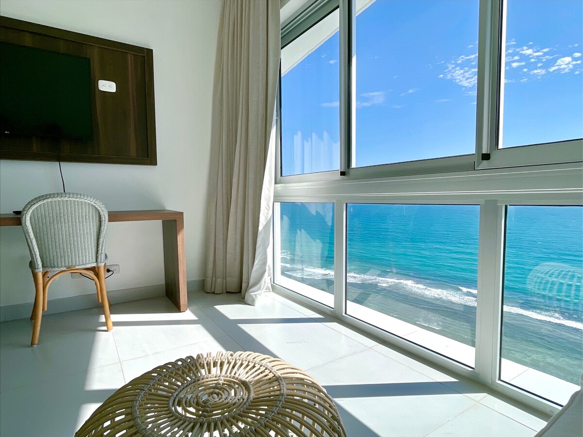 A bright corner of a living space is illuminated by natural light streaming through large floor-to-ceiling windows. The view features an expansive ocean, with gentle waves visible. A light wood desk and a comfortable chair are positioned to enjoy the scenery, enhancing a relaxed atmosphere.