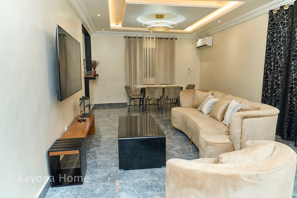 The inviting living area features a curved beige sofa arranged around a sleek black coffee table. A flat-screen television is mounted on the wall. A dining table with upholstered chairs is visible in the background, complemented by floor-to-ceiling curtains on the windows.