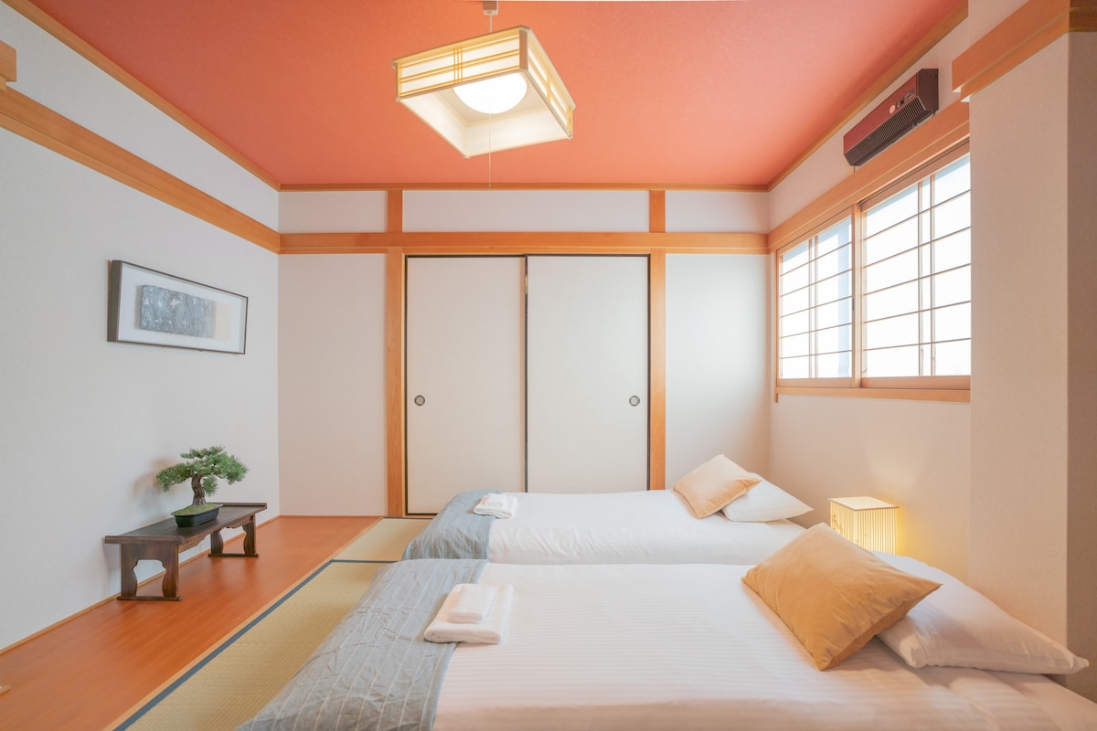 A serene bedroom features two spacious beds with neutral linens and decorative pillows. Natural light filters through window panes, illuminating the light-colored walls. A small potted plant decorates a low table, and a modern ceiling light adds gentle illumination.