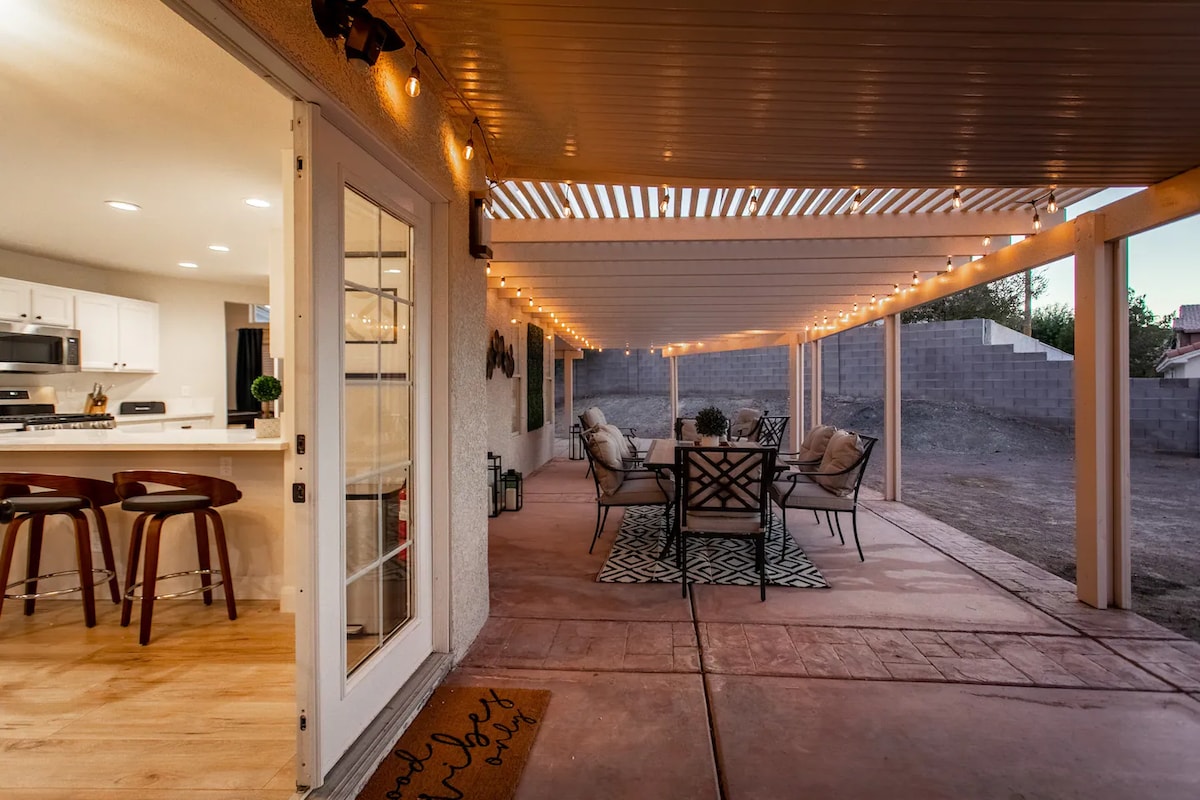 An inviting covered patio area connects to the indoor kitchen, featuring a seating arrangement with comfortable chairs and a decorative rug. Soft lighting is provided by string lights overhead, enhancing the space for relaxation and gatherings.