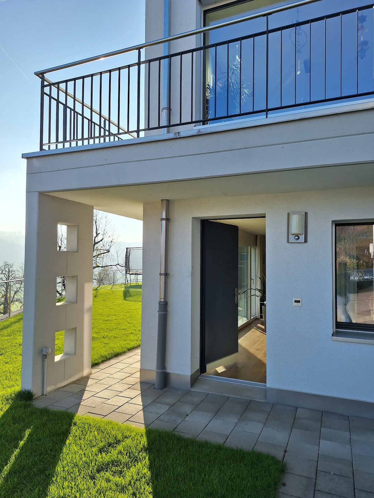 The exterior of the accommodation features a private entrance with a solid door, positioned at the base of a white building. Sunlight casts shadows across the stone pathway and well-maintained green grass, enhancing the inviting entranceway.