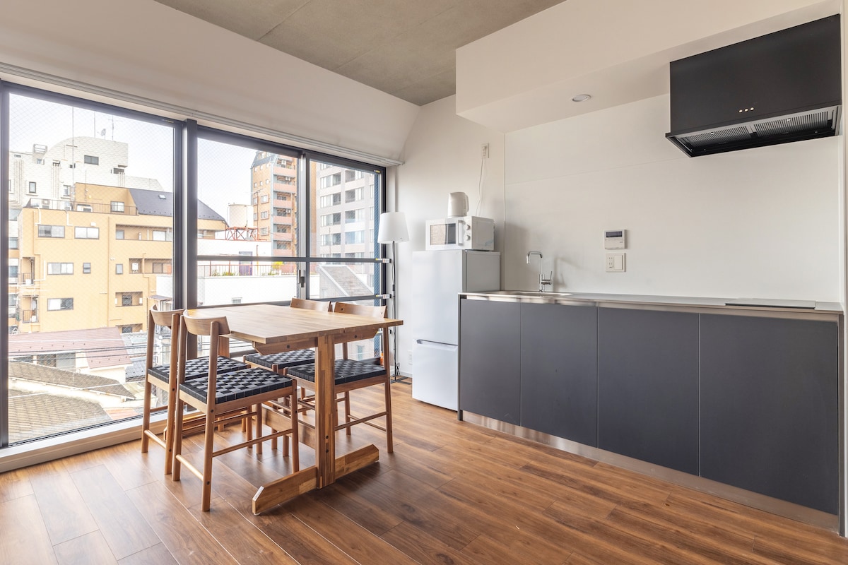 A modern kitchen area features sleek cabinetry and a stainless-steel refrigerator. A dining table with wooden chairs is positioned by large windows, allowing natural light to fill the space. The countertop includes a microwave and overhead range hood, creating a functional cooking environment.