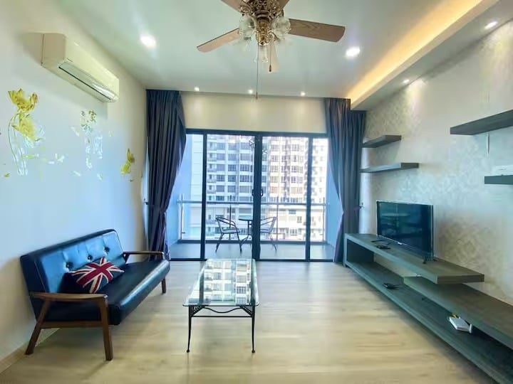 A modern living area features a sleek black sofa and a glass coffee table, complemented by light wood flooring. Large glass doors open to a balcony, allowing natural light to fill the space. Decorative shelves line the wall, enhancing the contemporary aesthetic.