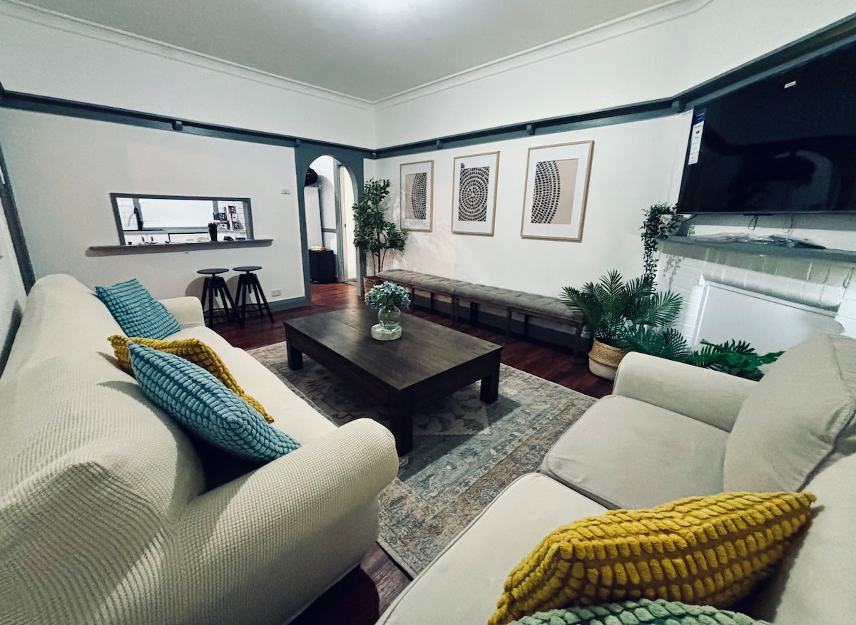 A comfortable living room is highlighted by a light-colored sofa with textured cushions and a dark wooden coffee table. Wall art pieces adorn the walls, while greenery adds a natural touch. A large television and an open doorway contribute to a spacious feel.