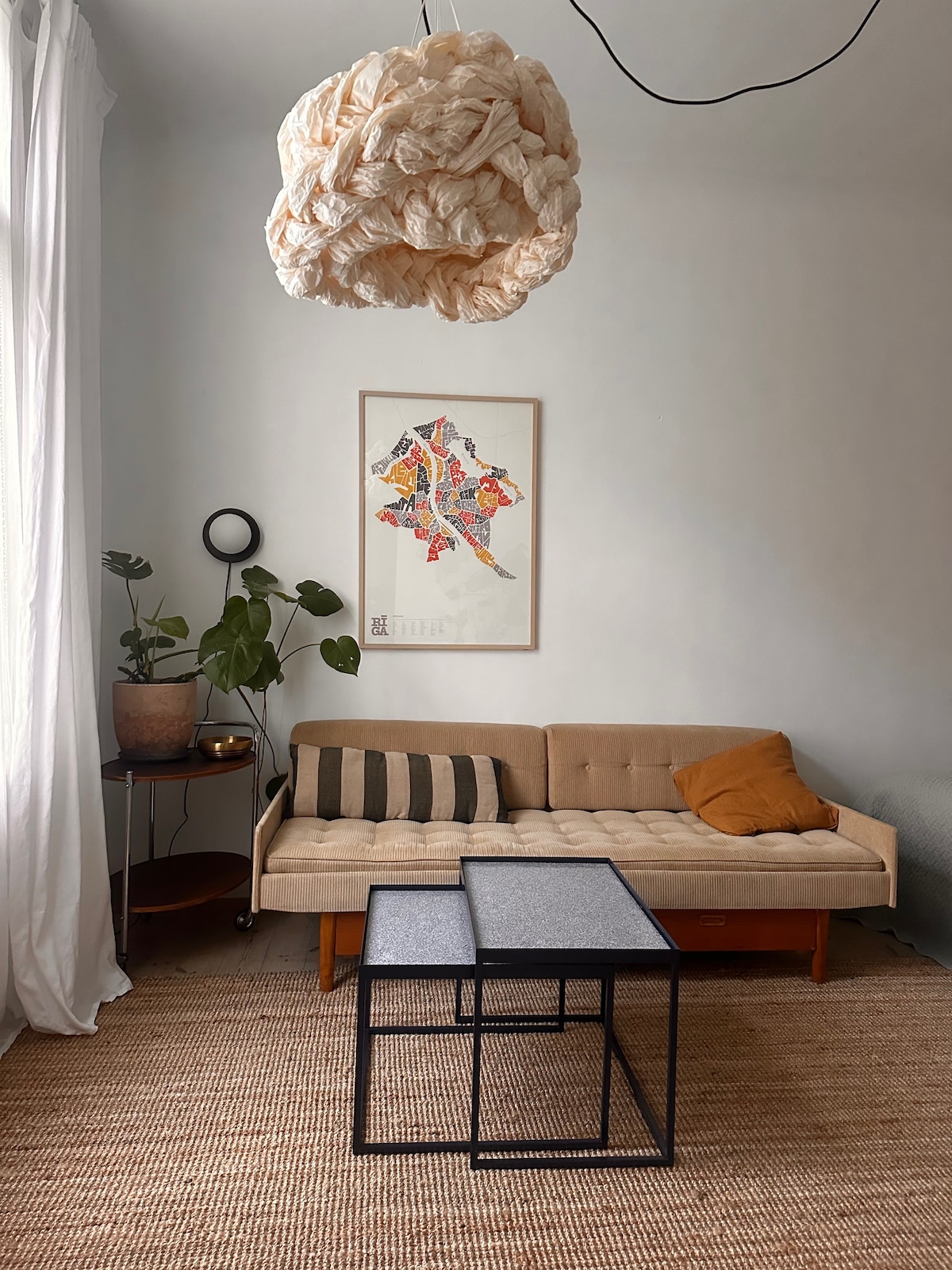A light-filled living area features a beige sofa with decorative pillows, set against a soft gray wall. Two modern coffee tables are positioned in front, while a colorful wall art piece hangs above. A potted plant and a floor lamp add natural and ambient elements.