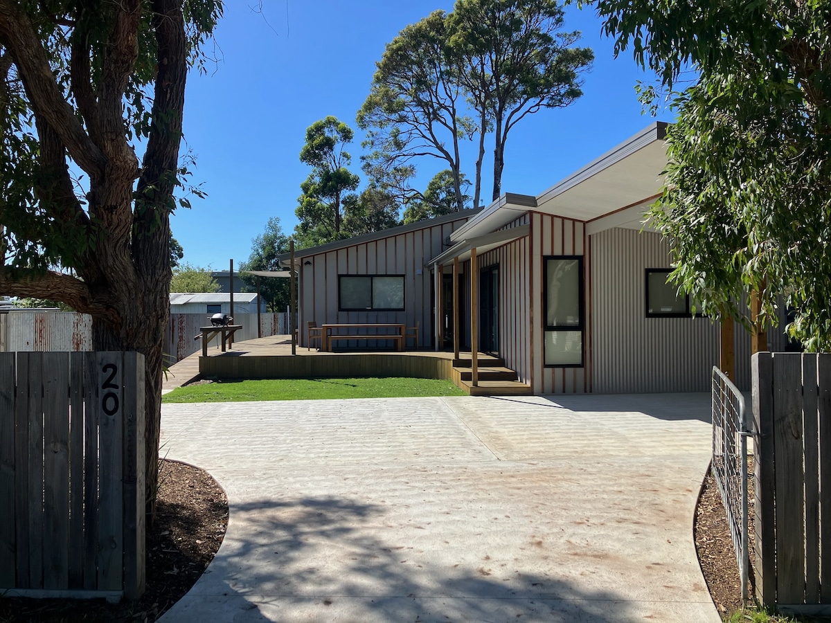 A modern, single-story home with a unique design features a spacious driveway in front, surrounded by landscaped greenery. Large windows enhance natural light. An outdoor seating area is visible, providing a welcoming entryway. The property is set within a serene environment with trees nearby.