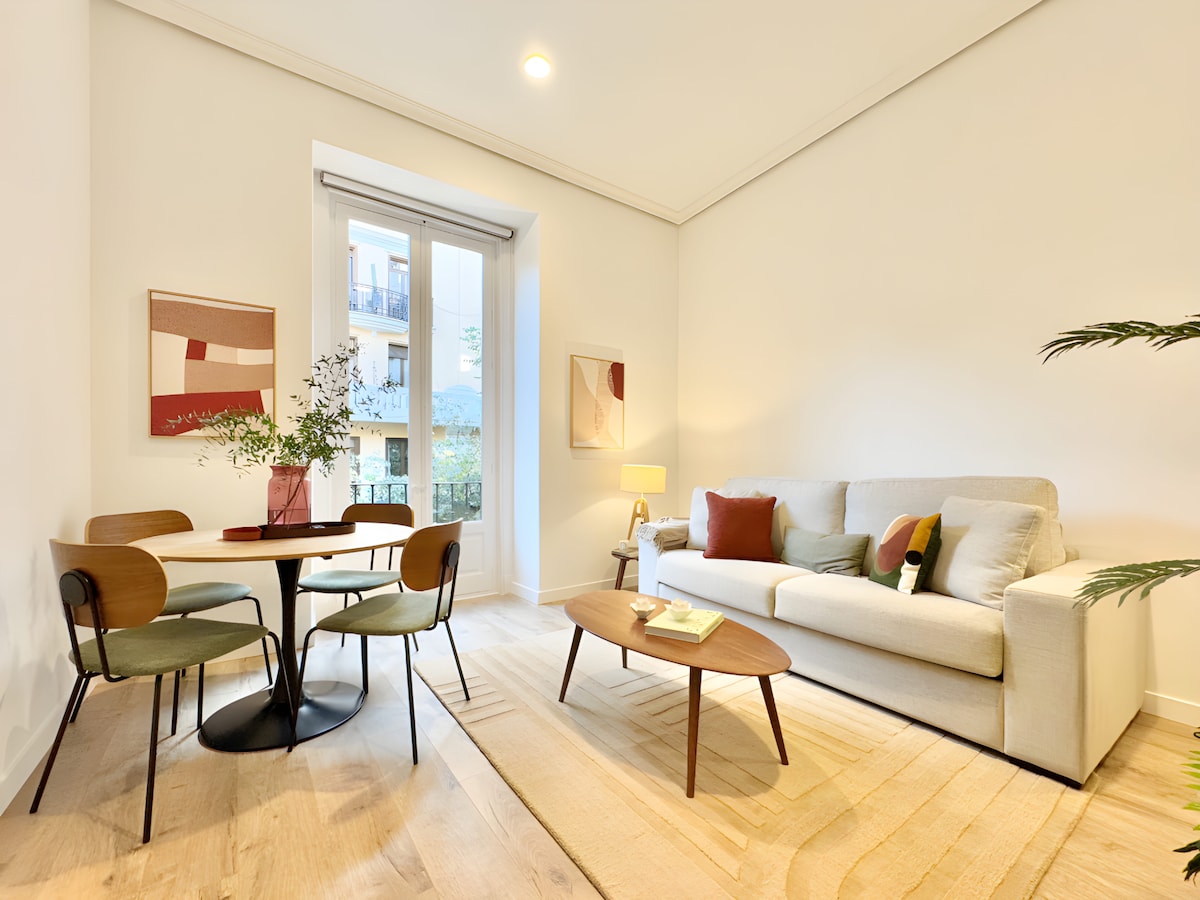 A bright and airy living room features a comfortable sofa, a circular dining table with chairs, and a decorative coffee table. Floor-to-ceiling windows bathe the space in natural light, offering views of the balcony. Minimalist artworks adorn the walls, complementing the modern design.