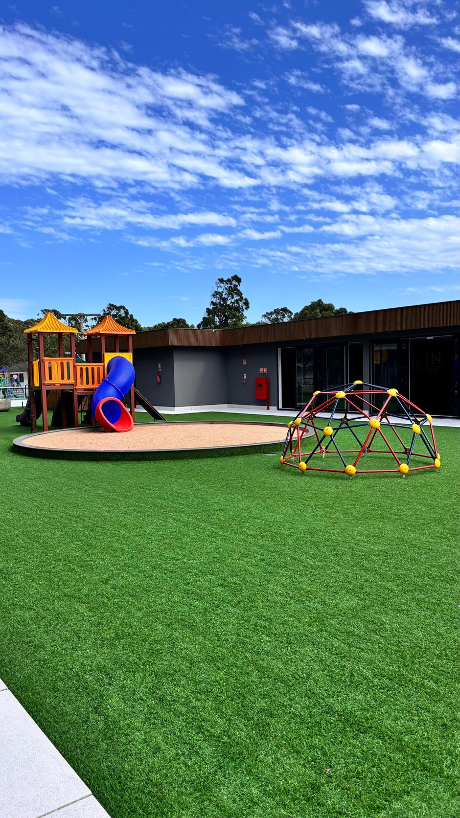 An outdoor play area is depicted, featuring a wooden play structure with a slide and a sandy base. A colorful climbing dome is positioned on the artificial grass, while blue skies and scattered clouds provide a bright backdrop. The modern building is visible in the background.