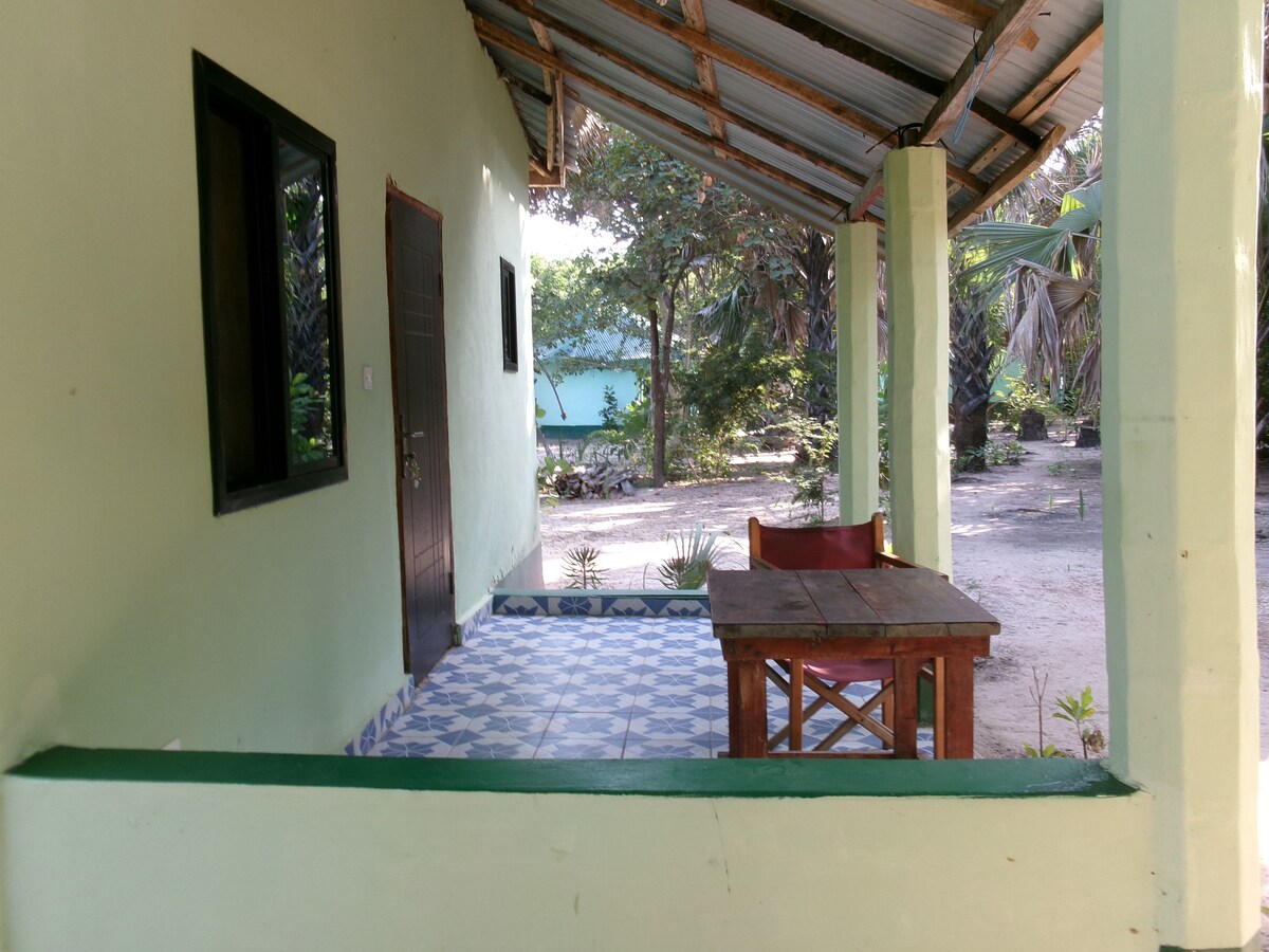 An outdoor seating area is showcased with a wooden table and red chairs, offering a place to relax. The surrounding landscape features tropical vegetation and a glimpse of the beach in the background, all under a partially shaded roof.