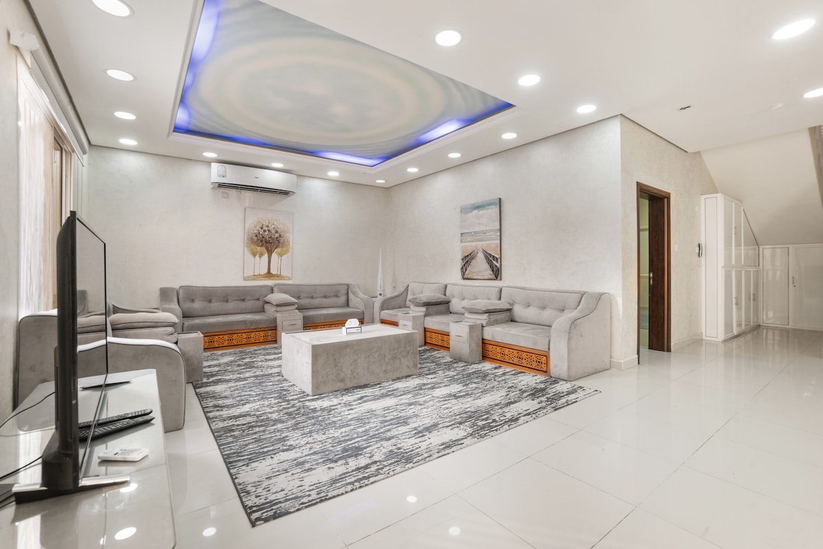 A spacious living area features plush grey sofas arranged around a central coffee table on a patterned rug. A subtle textured ceiling with ambient lighting enhances the room's feel. Natural light filters through large windows, complemented by decorative artwork on the walls.