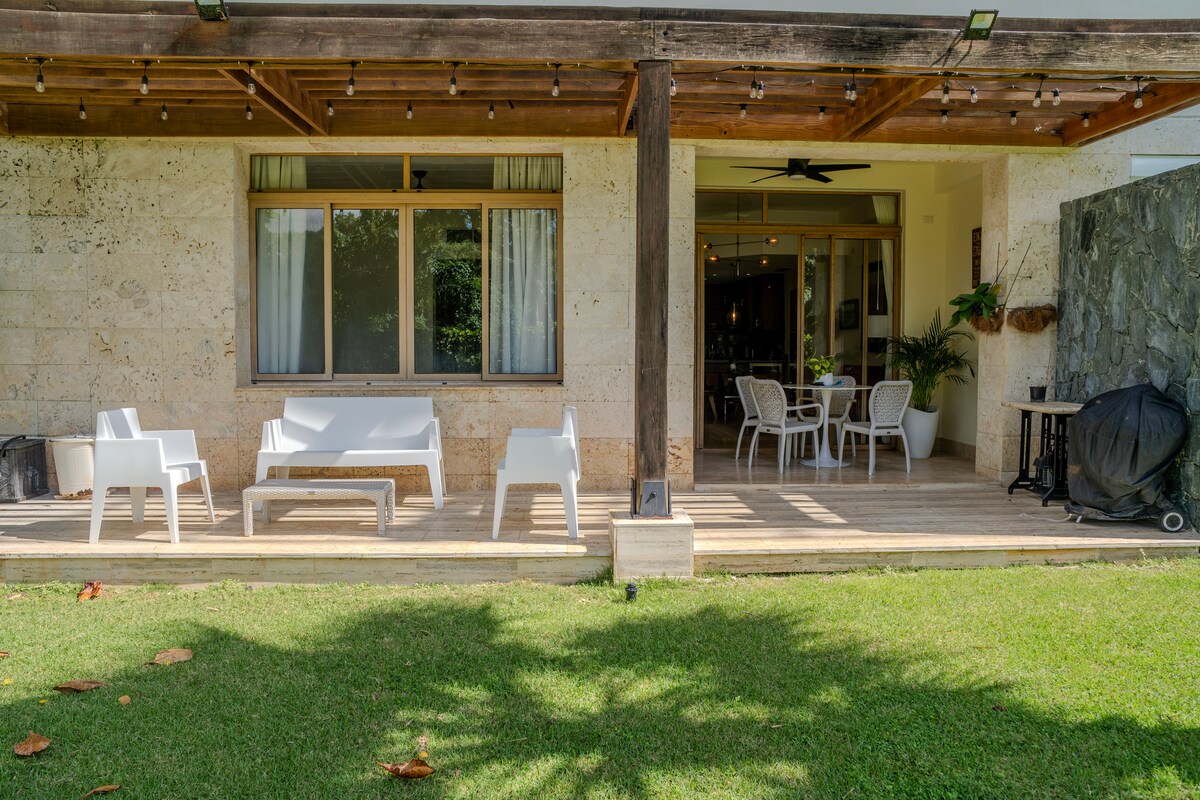 A covered outdoor patio is visible, featuring several white lounge chairs and a small black table. The natural wood beams and stone walls complement the green lawn, while large glass doors provide access to the indoor living space and dining area.