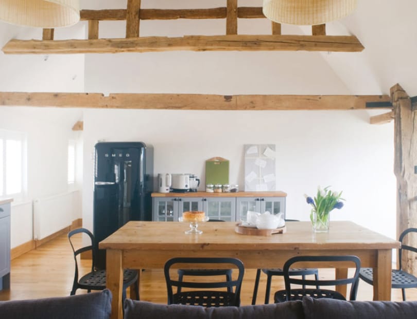 A spacious dining area features a large wooden table surrounded by six black chairs. A view of the kitchen showcases a black refrigerator, kitchen appliances, and minimalist decor. Natural light filters through multiple windows, enhancing the warm tones of the wooden beams overhead.