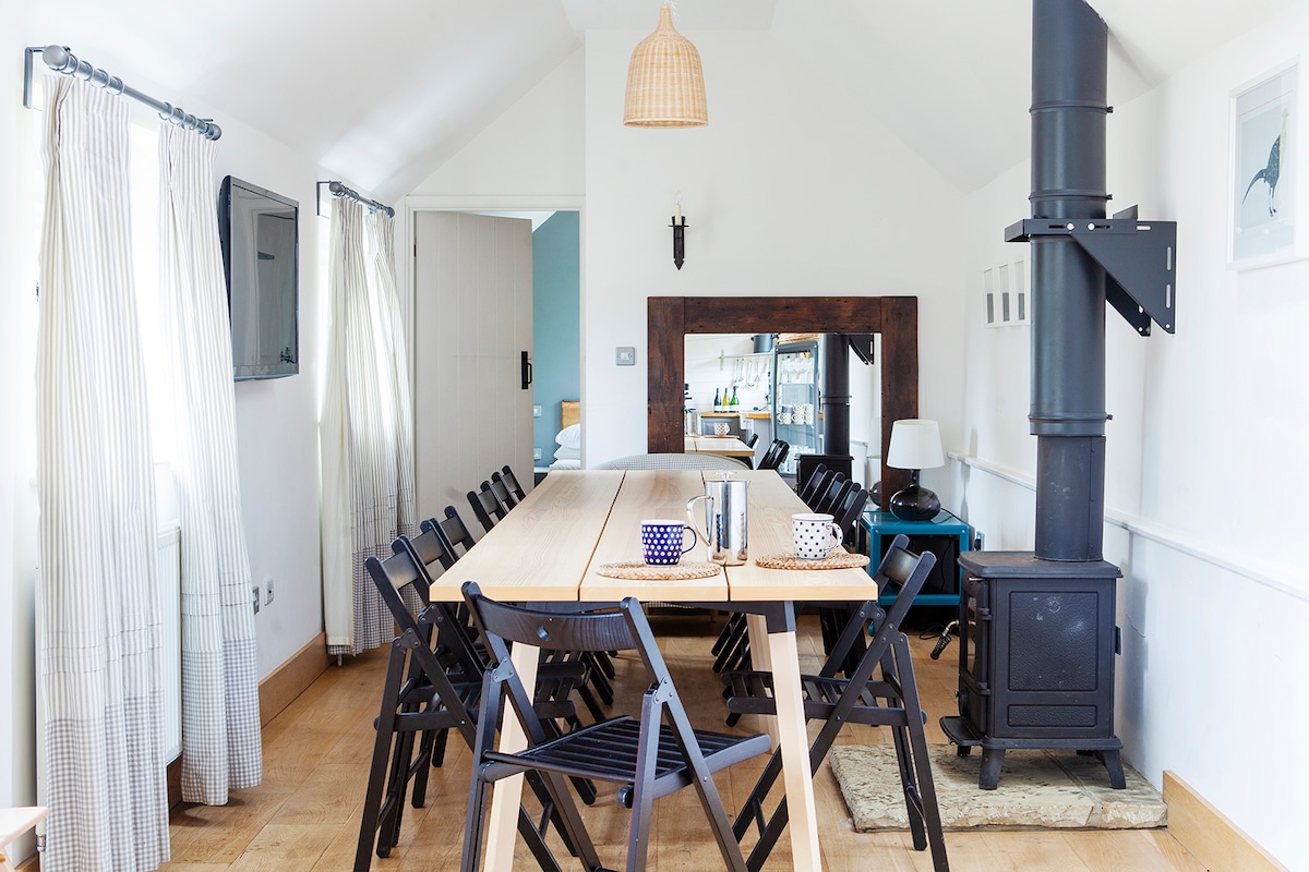 A spacious dining area features a long wooden table surrounded by multiple black folding chairs. A wood-burning stove stands in the corner, and a mirror reflects natural light from the nearby window. Light-colored curtains frame the window, complementing the modern decor.