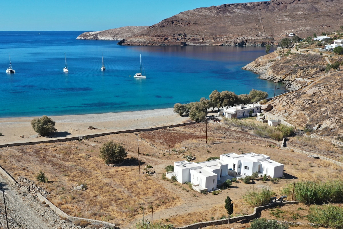 A scenic view of Ganema Beach is presented, featuring sand and calm blue waters with several sailboats anchored nearby. The surrounding landscape consists of dry earth and sparse vegetation, with white villas positioned on the beach, framed by a backdrop of rocky hills.