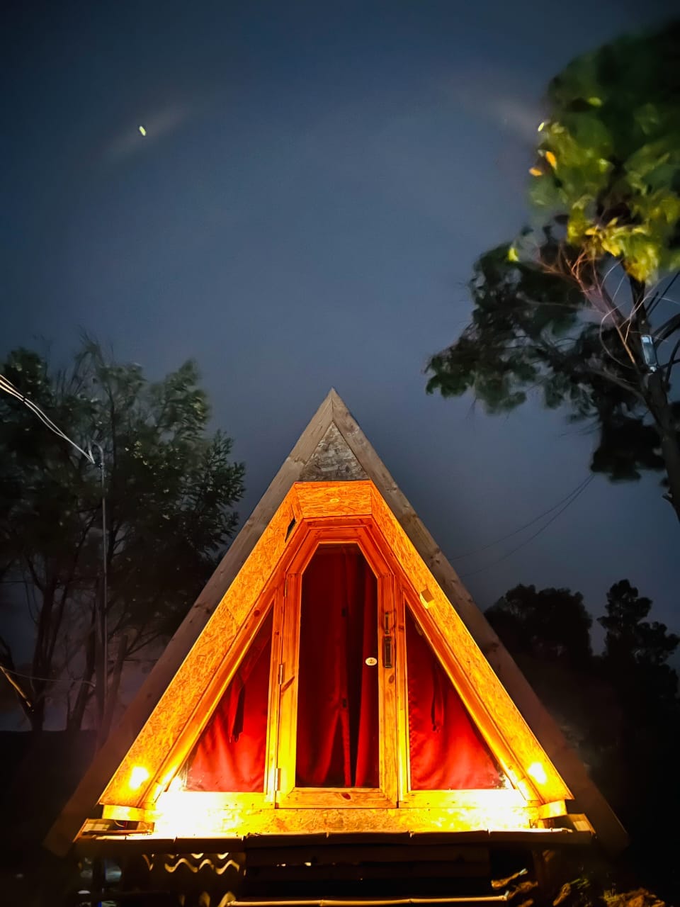An A-frame structure is showcased under a night sky, illuminated by warm exterior lighting. Red curtains frame the large windows, enhancing the unique triangular design. The surrounding trees add a natural element, contrasting with the geometric lines of the cabin.