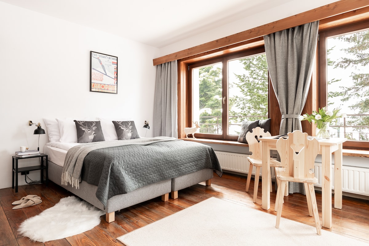 A well-lit bedroom features a spacious bed dressed in neutral tones, complemented by decorative pillows. Natural light floods through large windows, which are draped with soft grey curtains. A small wooden dining table with unique chair designs and a cozy rug lie adjacent to the bed.