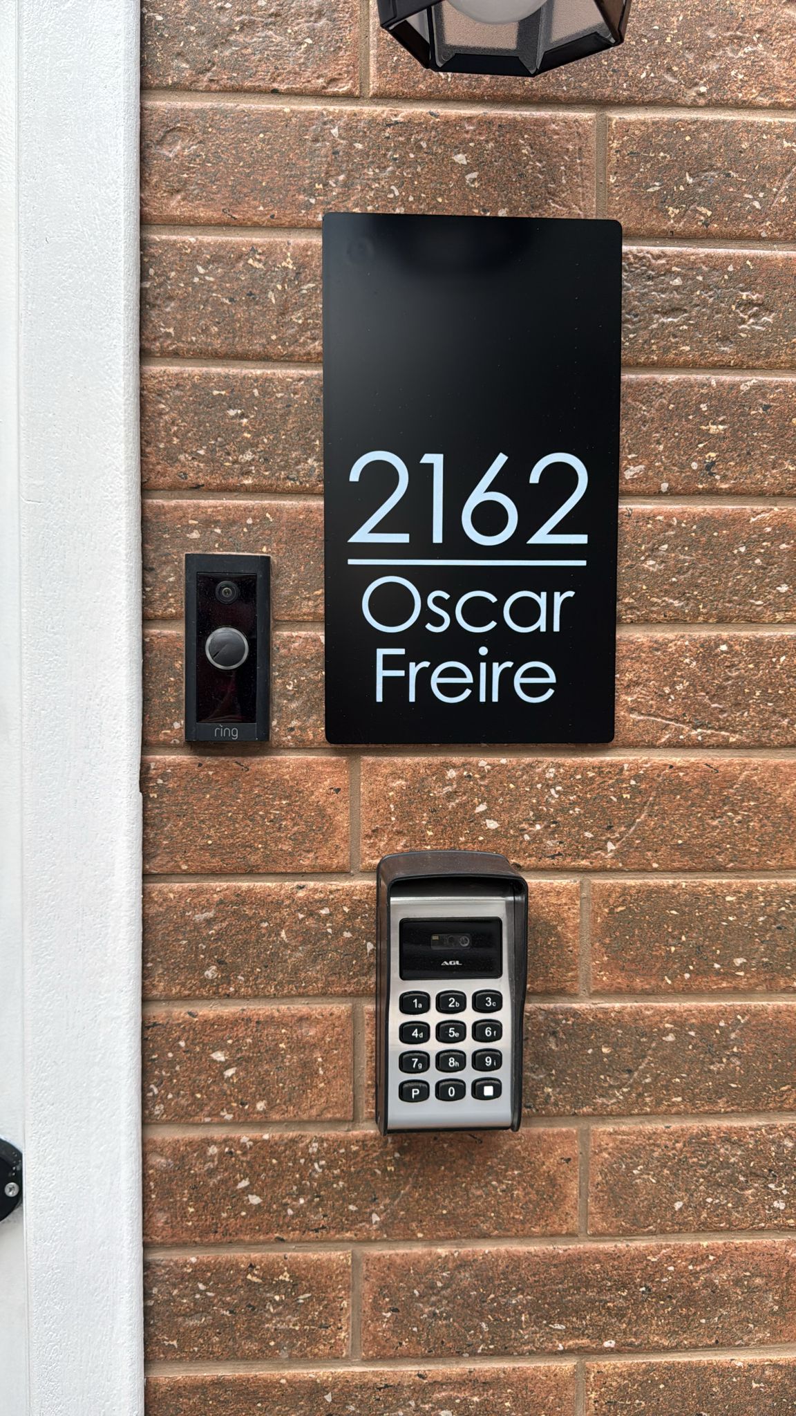 The entrance features a numbered black sign displaying '2162 Oscar Freire.' A digital keypad for access is positioned beneath the sign, alongside a doorbell intercom. The brick wall provides a textured background, enhancing the modern appearance of the entryway.
