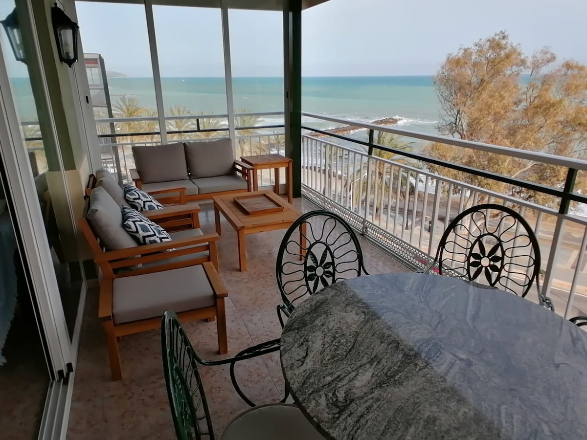 A terrace area is presented with stylish outdoor furniture, including a circular table surrounded by four chairs. Two cozy sofas with patterned cushions face the sea. The expansive view of the ocean and coastline is visible through glass railing, enhancing the inviting atmosphere.