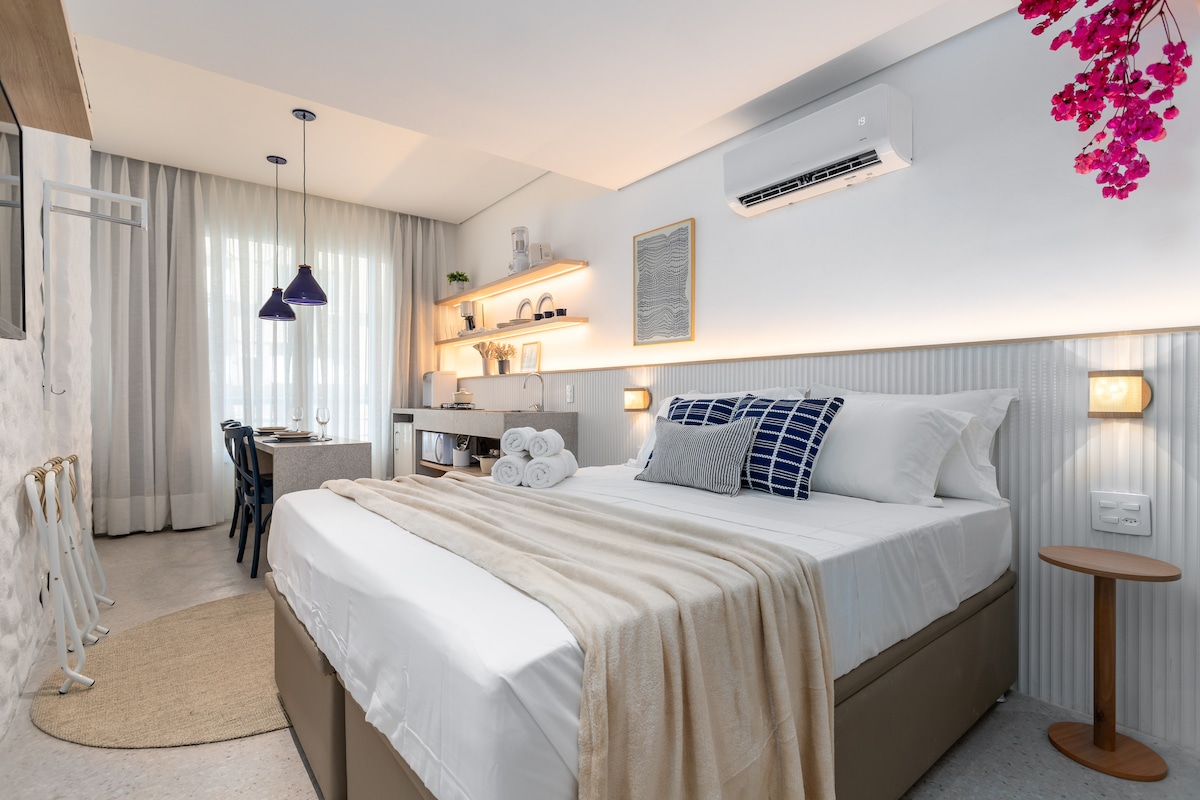 A modern studio features a spacious bed adorned with neutral linens and decorative pillows. Natural light fills the space through large, sheer curtains. The kitchenette is equipped with essential appliances, including a table suitable for dining, illuminated by stylish pendant lights.