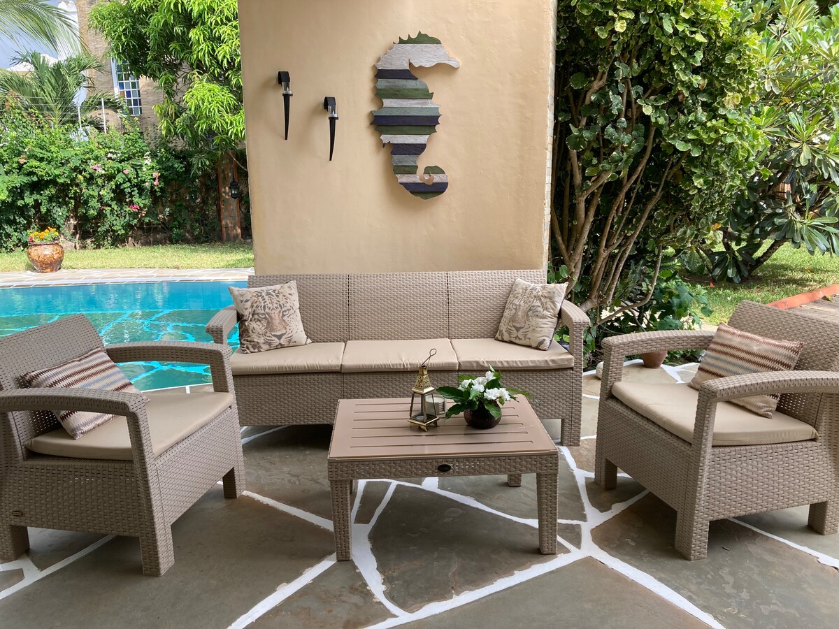 An outdoor seating area features a woven rattan sofa and two armchairs, arranged around a low coffee table. Decorative cushions add comfort. A turquoise pool is visible in the background, surrounded by lush greenery, contributing to a relaxing atmosphere.