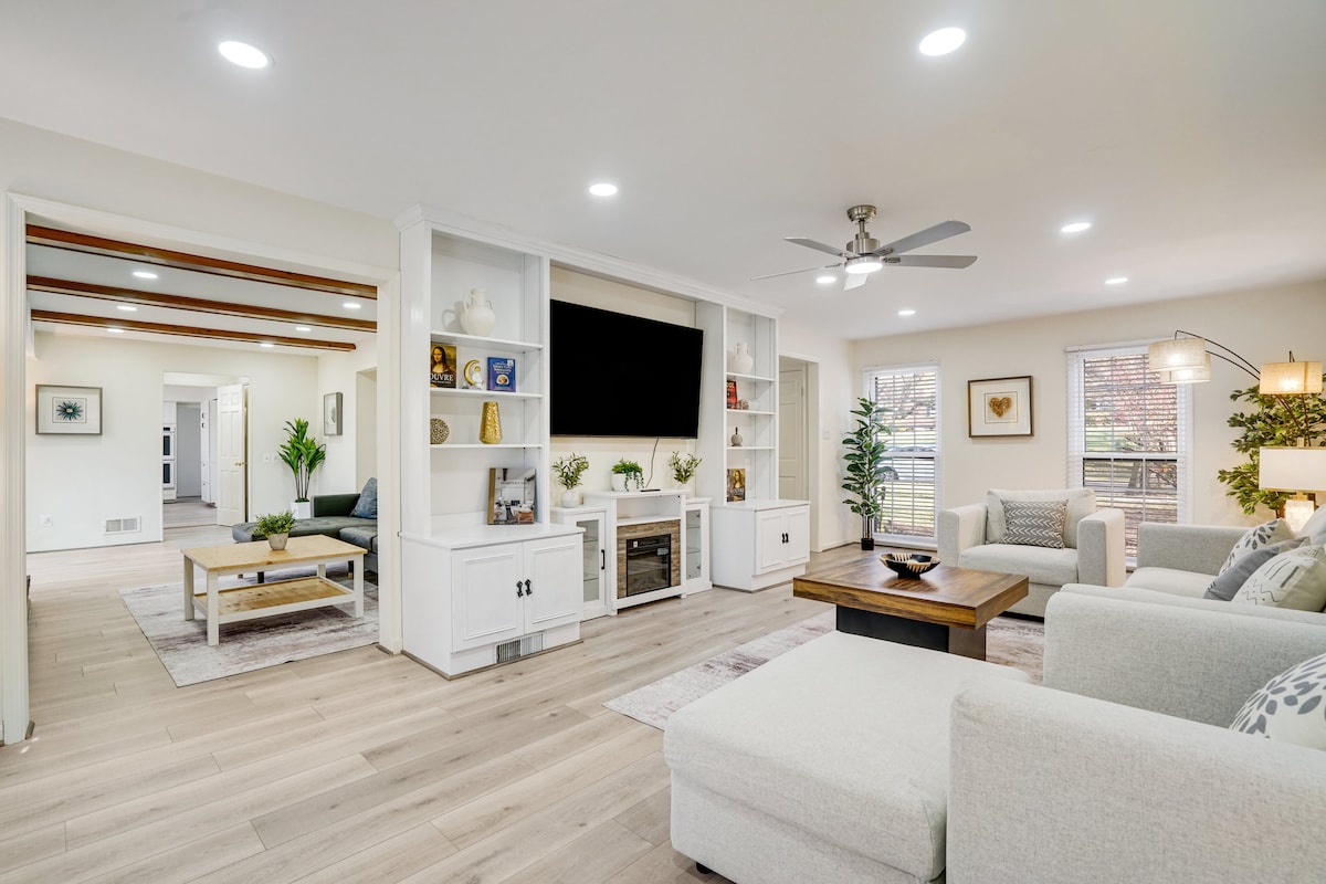 A spacious living area displays a cozy sectional sofa and a comfortable armchair, with a wooden coffee table at the center. A large TV is mounted on the wall, surrounded by built-in shelves. Natural light pours in through windows, creating a bright atmosphere.