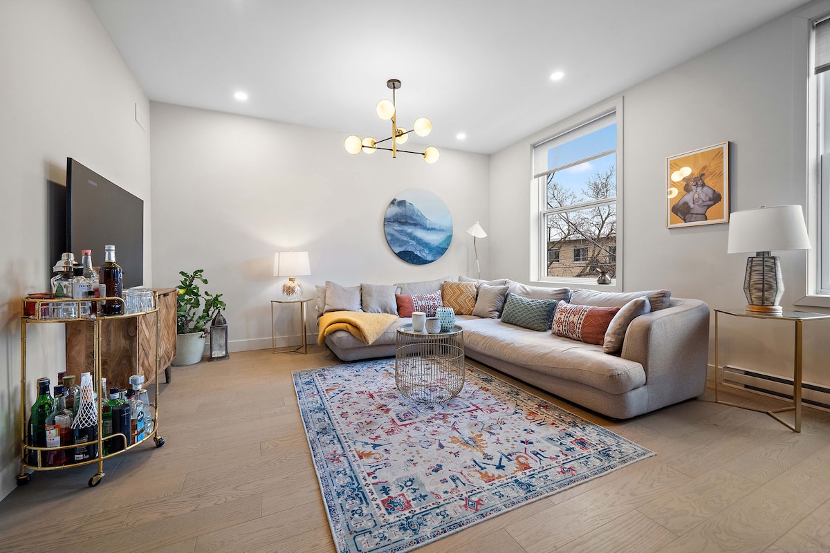 A bright living area features a spacious sectional sofa adorned with colorful cushions. An abstract wall piece complements the room, while a glass coffee table is centrally positioned on a patterned rug. Natural light enters through the large windows, enhancing the welcoming ambiance.
