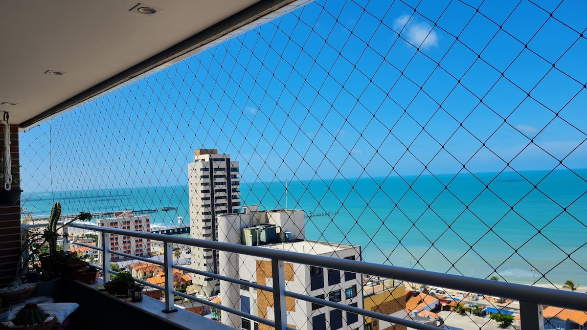A balcony view showcases a blue ocean under a clear sky, framed by a protective netting. Buildings of varying heights are visible along the shoreline, providing a sense of the vibrant coastal environment. Potted plants add a touch of greenery to the balcony space.