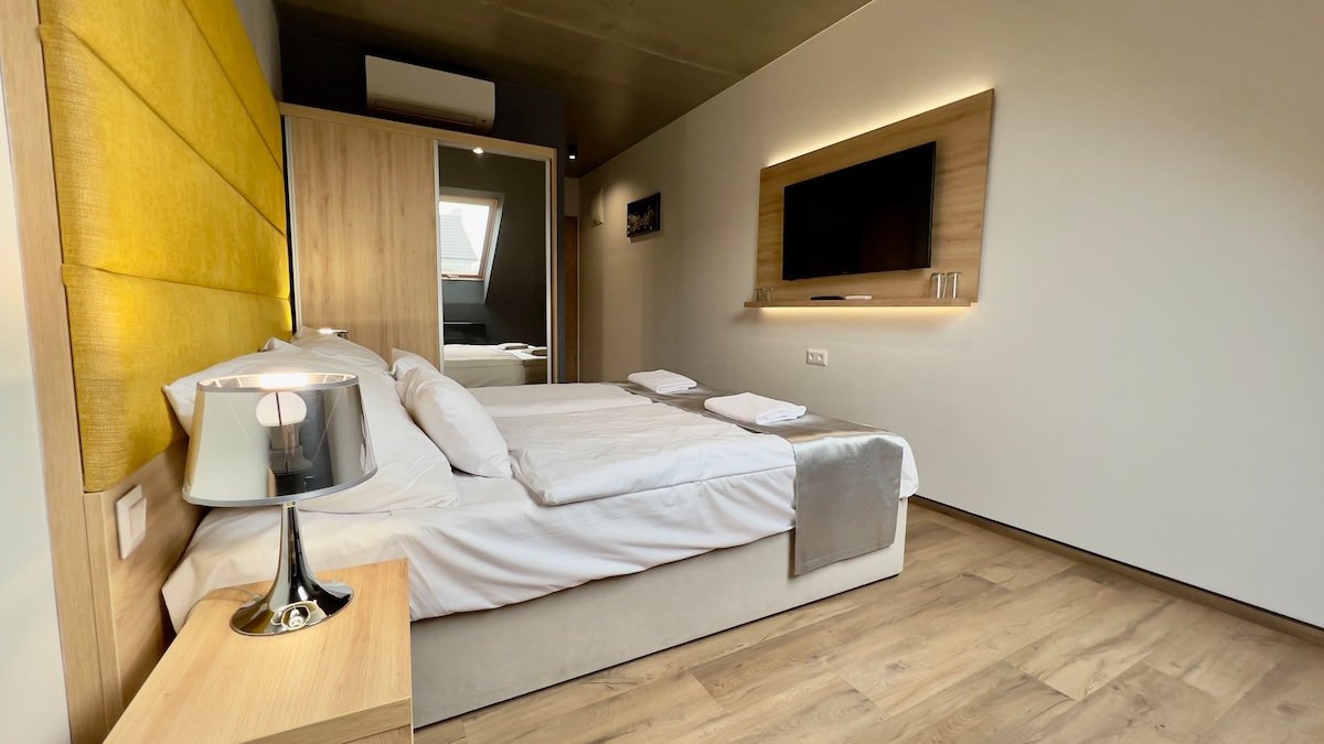 A modern bedroom features a spacious bed with white linens and plush pillows. A sleek bedside lamp sits on a wooden nightstand. The room is enhanced by warm yellow accent walls and a flat-screen TV mounted on the wall, creating a cozy yet contemporary atmosphere.
